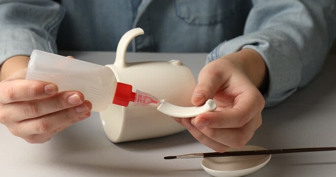 Woman gluing together pieces of broken mug at light grey table, closeup
