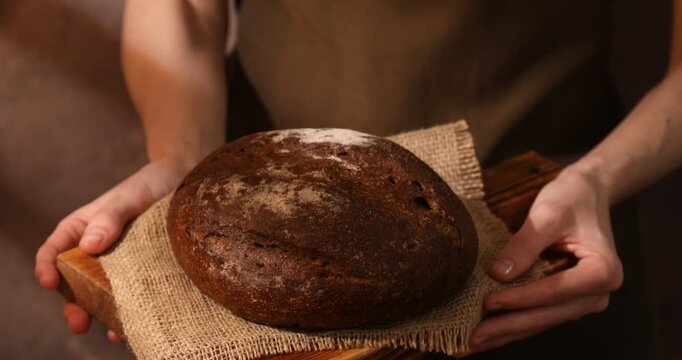 Woman holding loaf of fresh rye bread on brown background, closeup