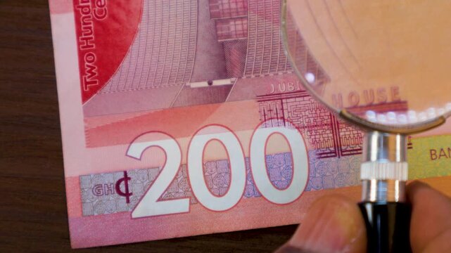 A magnifying glass inspects a 200 Ghanaian Cedi note, zooming in on the Jubilee House image, the denomination, and security symbols. The detailed scan highlights the bill&rsquo;s color and texture.