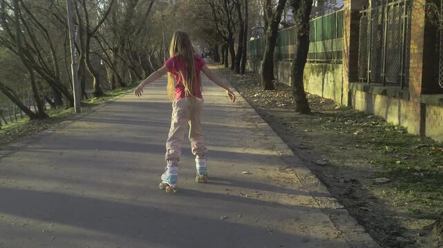 An editorial, tracking slow-motion shot following a young girl in a red shirt as she glides smoothly on roller skates down a paved walkway lined with trees and a fence in Budapest during the late afte