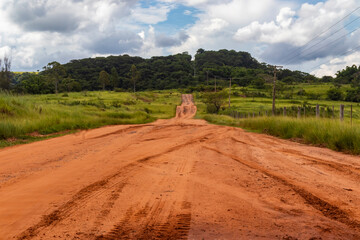 Naklejka premium Red dirt rural road crossing farmland under dramatic storm clouds, with power lines and a lone dry tree, depicting countryside landscape, severe weather and agricultural infrastructure.
