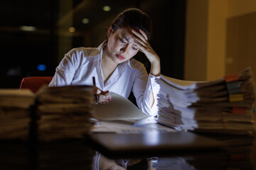 A woman is seen studying papers in a dark office, showcasing stress and focus while surrounded by piles of documents, emphasizing the pressure of her workload and late-night efforts. © David