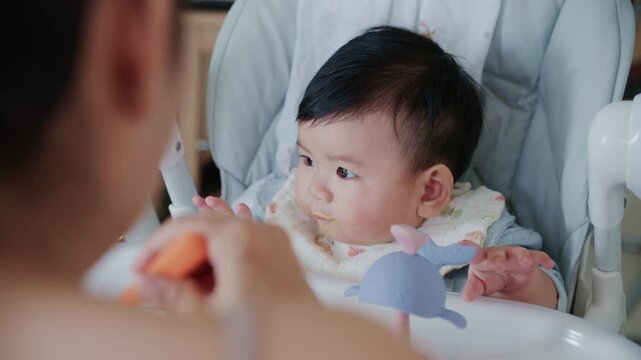 4K Asian mother feeding her six month old baby boy with a spoon at home in morning.
