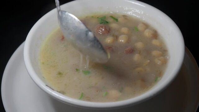 Close-up shot of a spoon stirring a bowl of homemade chickpea and sausage soup with fresh herbs, showcasing the creamy texture and delicious ingredients