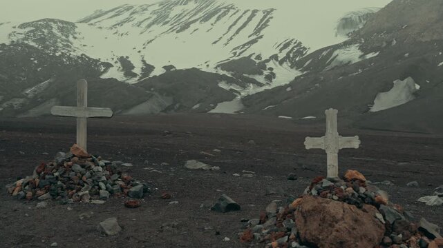 Two weathered crosses mark historic graves at Whalers Bay cemetery on Deception Island, Antarctica, surrounded by barren volcanic terrain and snow-patched mountains under gloomy skies
