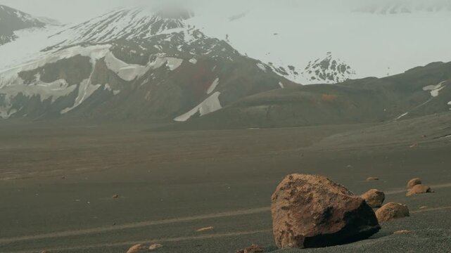Barren volcanic plain with rugged boulder stretches toward a snow-capped volcano at Whalers Bay, Deception Island, Antarctica, under hazy overcast skies
