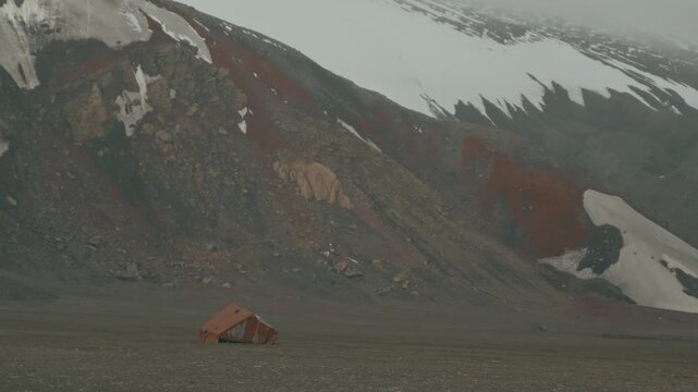 Rusted oil tank relic from historic whaling era sits alone on vast dark volcanic plain at Whalers Bay, Deception Island, dwarfed by dramatic rust-red snow-streaked slopes
