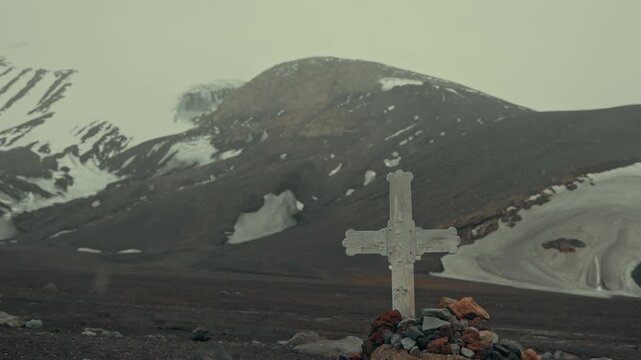 Weathered white cross on stone cairn marks historic whaler grave at Whalers Bay cemetery, Deception Island, with dark volcanic slopes and snow patches fading into misty sky