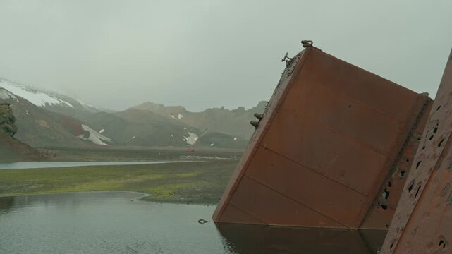 Massive rusted industrial tank from historic whaling station tilts into lagoon at Whalers Bay, Deception Island, Antarctica, with volcanic snow-patched mountains in background