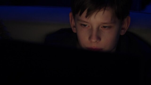 A teenager focuses on working at a laptop in a dark room late at night. The boy's face is illuminated by the flickering light from the screen against a blue interior backlight