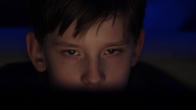Close-up portrait of a boy staring intently at a computer screen in a dark room. The child's face is illuminated by the soft light from the display against a blue neon background