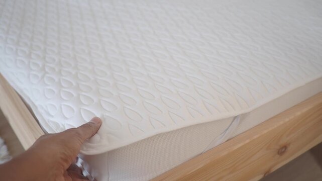 Close up of person hand adjusting white quilted mattress topper on wooden bed frame in bright bedroom