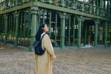 Fototapeta premium Woman with backpack in a park near ornate green architecture, outdoor stroll, coat and casual style, showcasing travel vibe, urban scenery and daylight calm.