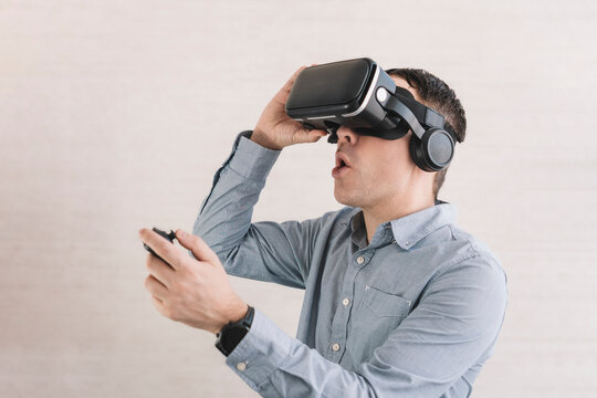 Young smiling man in VR glasses playing video game with joystick on a grey background, siting on the couch while having virtual reality experience, copy space