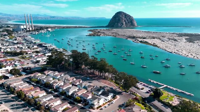 Aerial drone view of Morro Bay Harbor, Morro Rock, marina boats, coastal homes, and the former power plant on California&rsquo;s Central Coast under clear blue skies.
