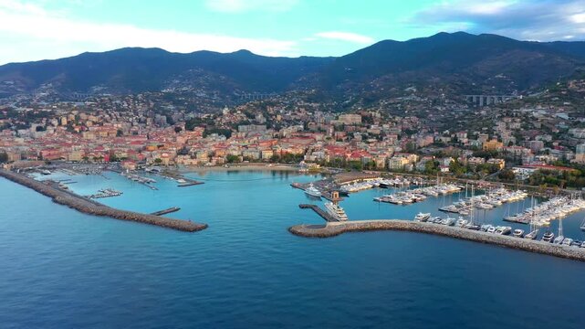 Drone view of San Remo harbour in Liguria, Italy, featuring luxury yachts, calm Mediterranean water, rocky breakwater, marina docks, and the hillside cityscape of the Italian Riviera.