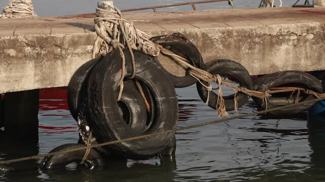 Old tires used as dock bumpers in Rayong. Close-up of ropes and maritime infrastructure with water reflections.