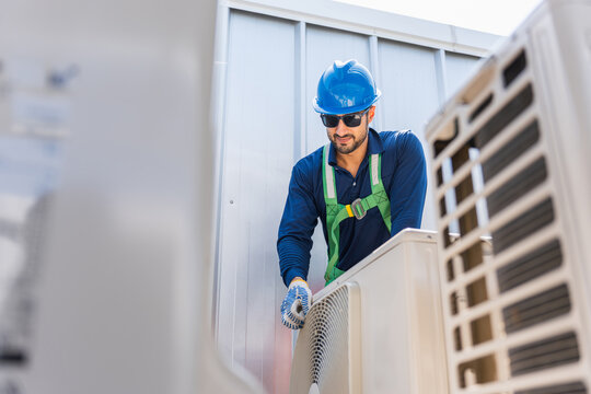 Professional HVAC technician in safety gear inspecting outdoor air conditioning unit on building rooftop, Technician performing routine maintenance service on external air conditioner compressor unit