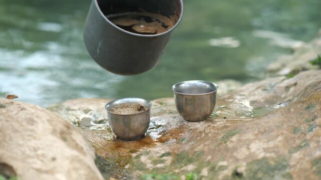 Close-up of coffee being poured into metal cups on rock near river.