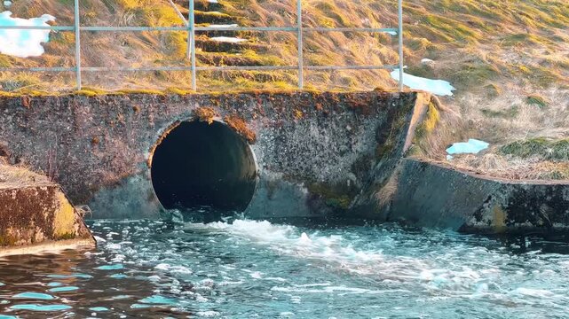 A concrete culvert pipe releases flowing water into a river. Suitable for media projects focused on urban infrastructure, environmental drainage systems, and wastewater management.