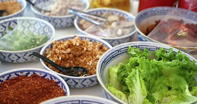 Assorted Thai noodle condiments on table including chili flakes, sugar, crushed peanuts, greens and soup, representing authentic Asian street food culture