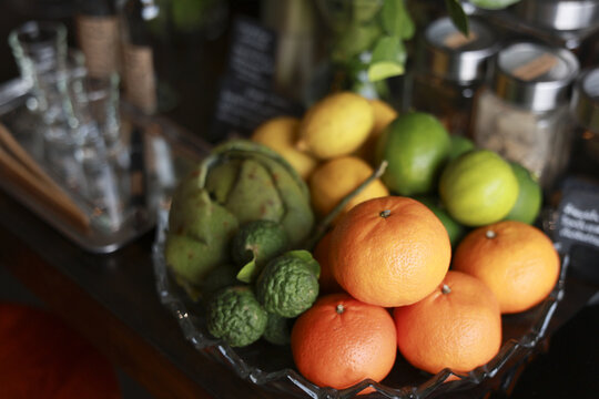Vibrant fresh citrus fruit arrangement featuring orange, lemon, lime, and kaffir lime in decorative glass bowl on bar counter, creating refreshing, natural, and inviting atmosphere