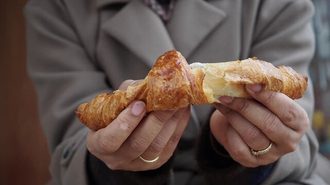 Close up video of person in neutral colored coat tearing golden baked croissant in half, showing flaky, airy texture inside