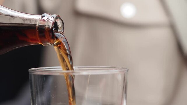 Close-up cinematic shot of dark carbonated drink, possibly soda or cola, being poured from glass bottle into clear glass, creating fizz and bubbles in a captivating slow motion video