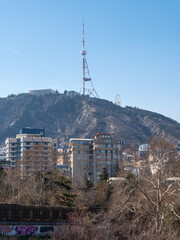 Daytime panoramic view of Tbilisi, Georgia, featuring the Mtkvari river, residential buildings, and the iconic TV broadcasting tower on top of Mtatsminda mountain under a clear blue sky. © Microscope