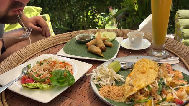 A person enjoys a delicious spread of authentic Thai dishes like Papaya salad, Pad Thai, and crispy spring rolls, served on a traditional rattan tray in a lush outdoor setting.