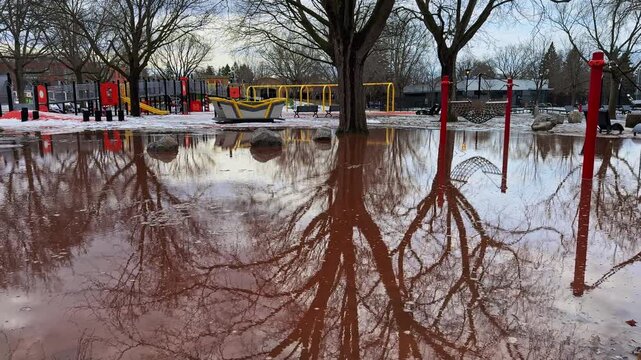 Reddish spring meltwater overtakes a public playground, where bare tree reflections and submerged play structures reveal a sudden, unusable park scene.