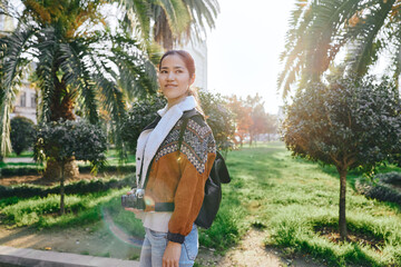 Fototapeta premium Woman with camera in sunny park, outdoor fashion, backpack, casual outfit, capturing moments amid palm trees and green lawn, bright warmth and relaxed mood