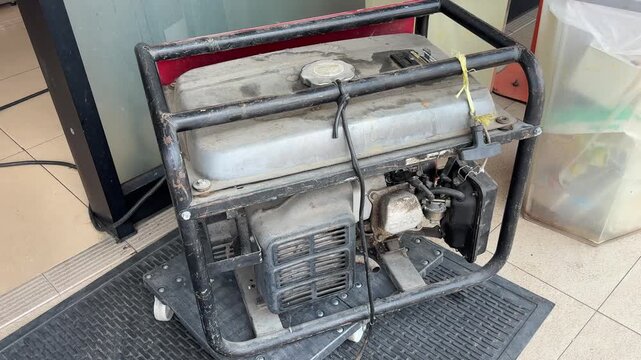 An old, dusty portable generator with a black frame sits on a wheeled platform, suggesting it's used for backup power. It appears to be located outdoors or in an open commercial area.