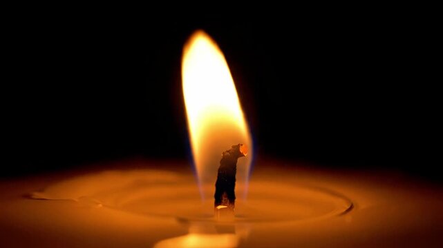 A warm, golden flame rises from a candle wick, casting a soft glow on the melted wax surface, set against a dark background