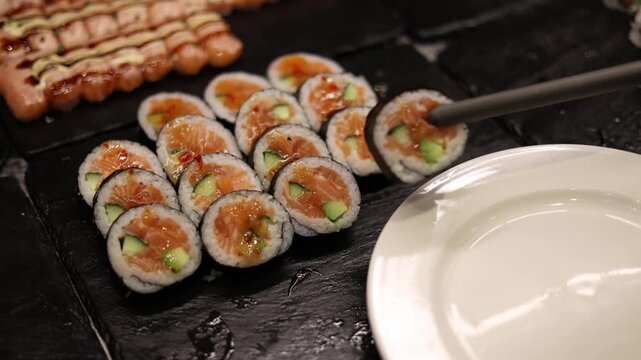 Sushi rolls with salmon being picked with tongs onto plate in restaurant showing serving process and fresh seafood selection for healthy meal