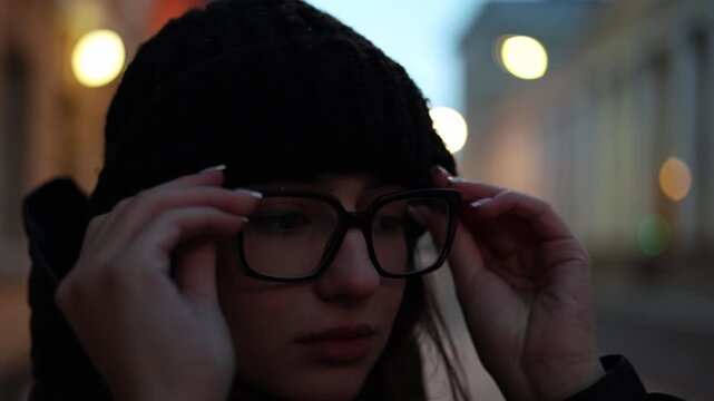 Young woman putting on eyeglasses and hood before walking into evening city creating cinematic lifestyle scene with urban atmosphere and calm mood