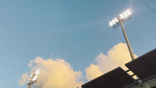 Low-angle view of a stadium floodlight tower glowing against a bright blue sky with drifting clouds, capturing a powerful sports atmosphere.