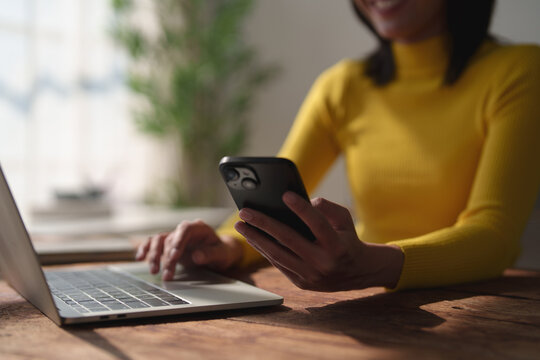 Woman with a yellow sweater sitting at a wooden desk at home, typing on a laptop keyboard and holding a smartphone, actively managing multiple digital tasks