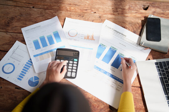 Person's hands working on a calculator and analyzing business loan comparison reports and financial graphs on a wooden desk, managing expenses and planning investments