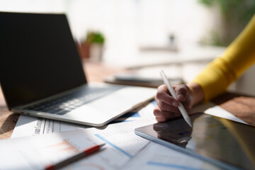 Fototapeta premium Business hand using a digital pen on a tablet screen, seeing charts and graphs, working on reports with a laptop and documents on a desk, representing modern finance and productivity