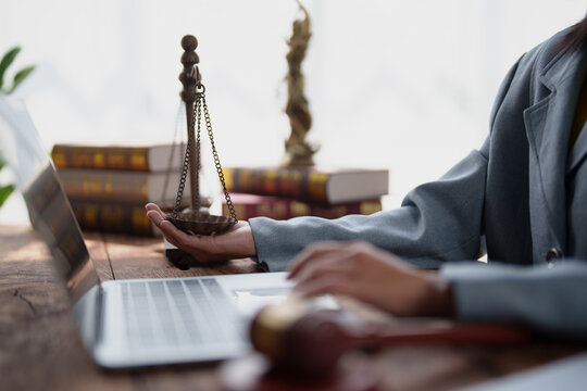 Legal professional's hand holding a justice scale, symbolizing law and equity, while typing on a laptop with legal books and a gavel in background, representing modern legal practice