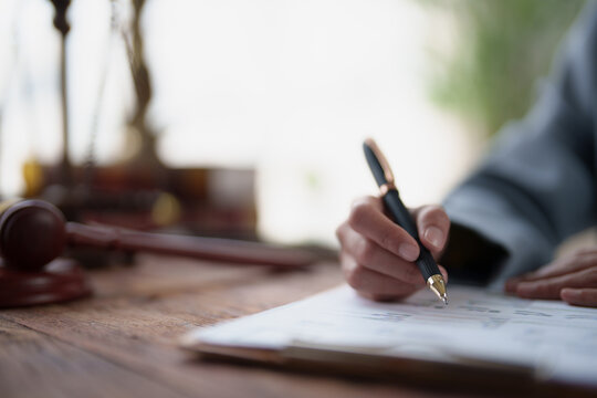 Lawyer hand writing on an important legal document or contract, with a blur of a gavel and scales of justice on a wooden desk, symbolizing the legal profession and business law