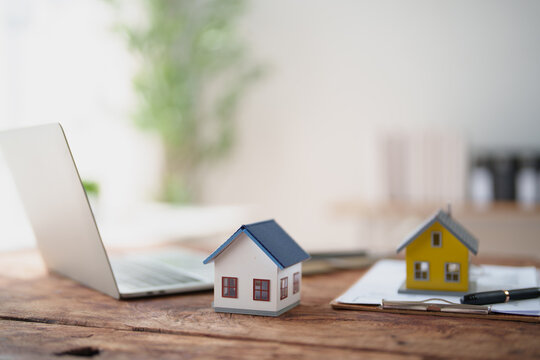 Two miniature houses on a rustic wooden desk beside an open laptop and clipboard with documents and pen, symbolizing real estate, mortgages, property investment and home buying decisions