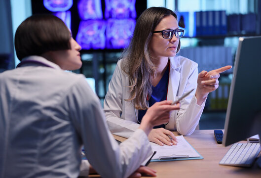 In a sleek, well-lit office, two doctors engage in a focused discussion about brain scans on a computer. One points at the screen, while the other takes notes, highlighting teamwork.