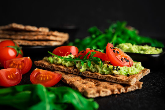 Avocado sandwich with avocado cream and rye crisp bread for snack. Fiber, fitness and diet food. Rye bread with guacamole, arugula and cherry tomatoes on dark background. High quality photo