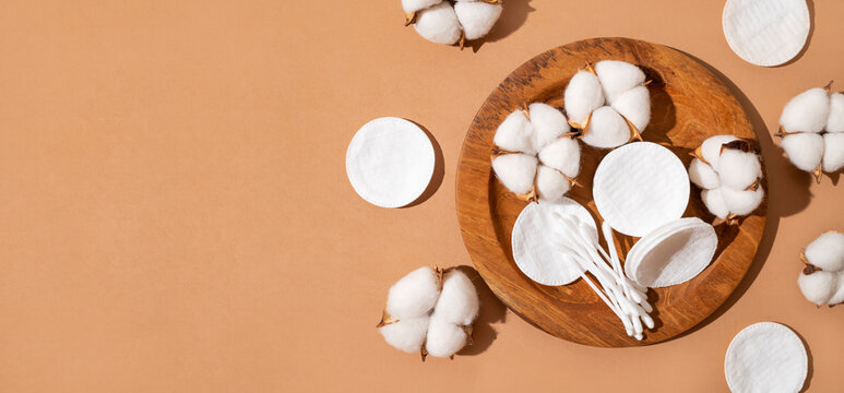 Natural cotton pads, cotton swabs and lush cotton flowers on wooden tray on brown background. Beauty, health and personal care concept. Cosmetic accessories for makeup removal and facial care