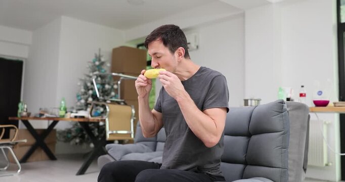Man eats corn on cob while sitting on sofa at home. Guy bites boiled kernels rotating cob while cleaning rows with steady chewing slow motion