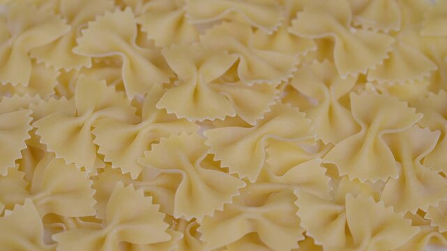 Closeup of dried bowtie pasta grains. Pile of warm beige uncooked ribbonshaped pasta slices. Detailed view of arranged dry farfalle pasta with serrated edges in warm tones