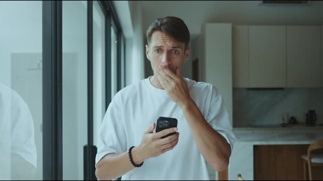 Man looks at smartphone in shock after seeing terrible news at home. This shock moment shows shock reaction stress and fear as shock emotion grows while reading alarming message on mobile phone