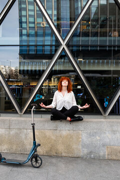Redhead meditating outdoors with scooter in front of modern city building
